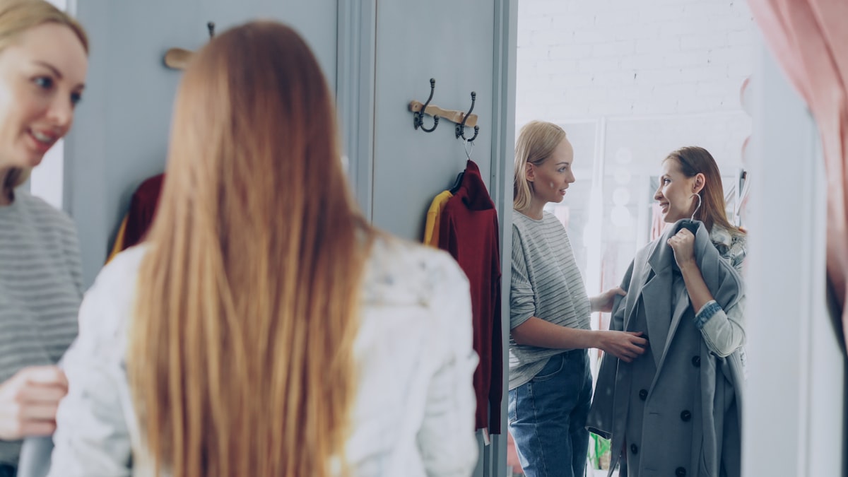 Two women discussing a coat in a fitting room mirror.